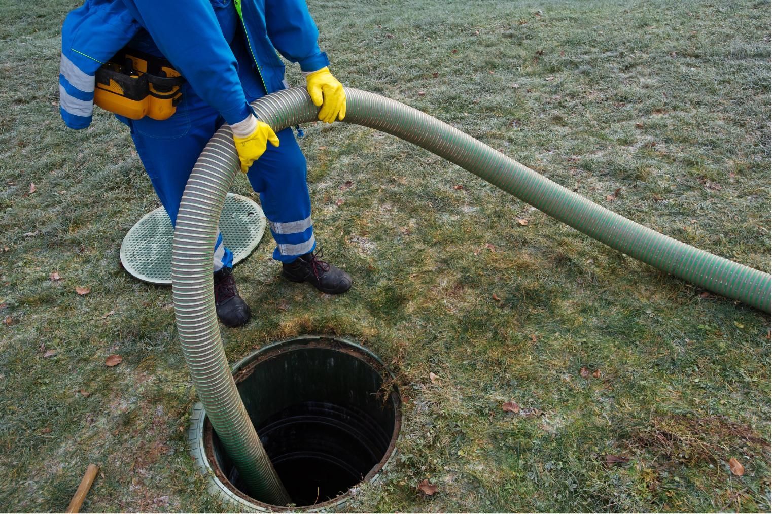 Worker in blue overalls and yellow gloves lowering a hose into an open manhole on frosty ground