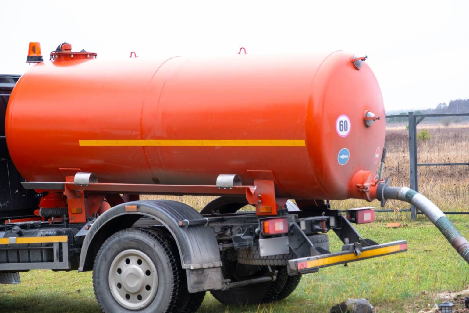 Orange fuel tanker trailer with hose attached, parked on a road near grassy fields