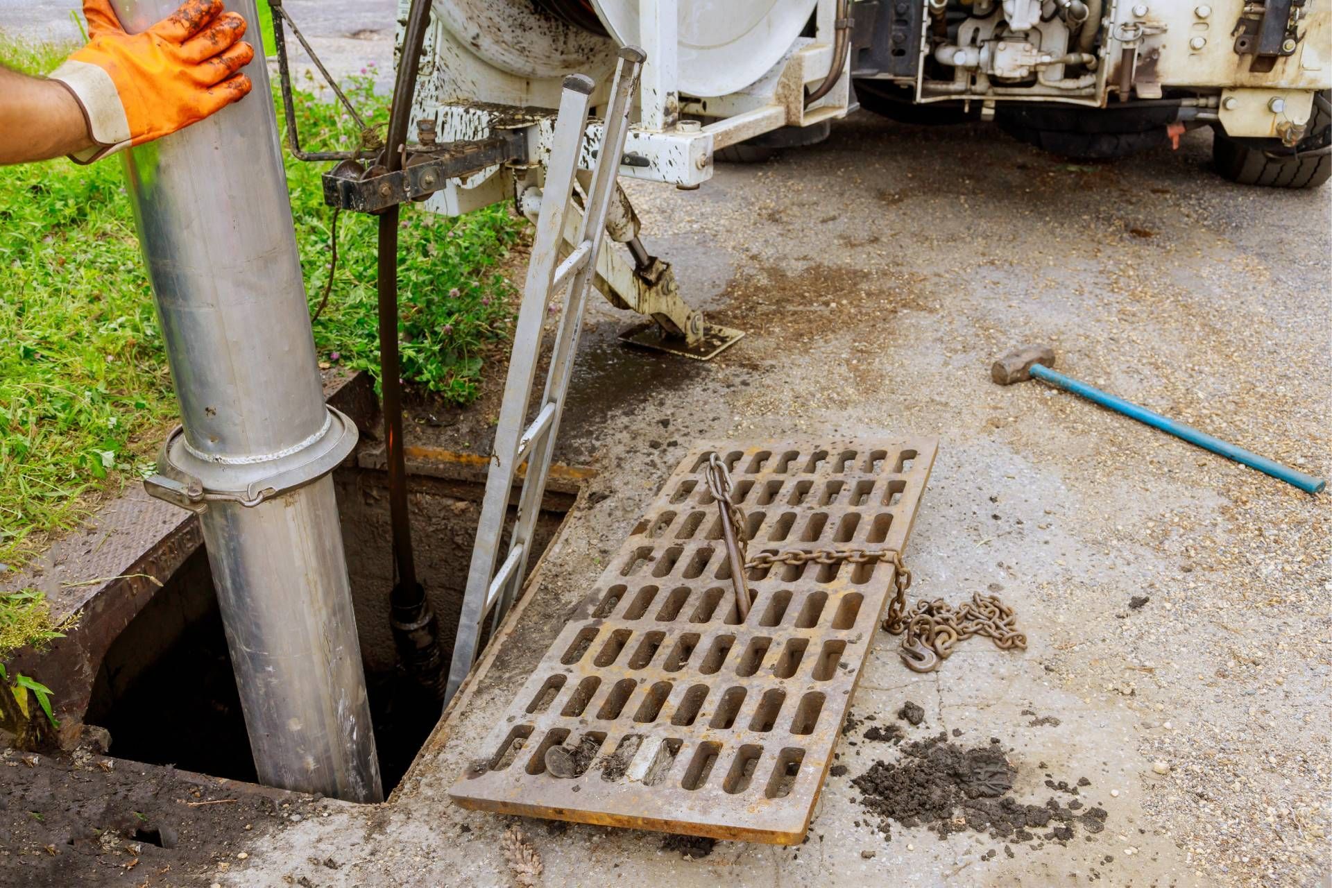 Gloved worker beside open manhole on road, with metal pipe and grate cover nearby.