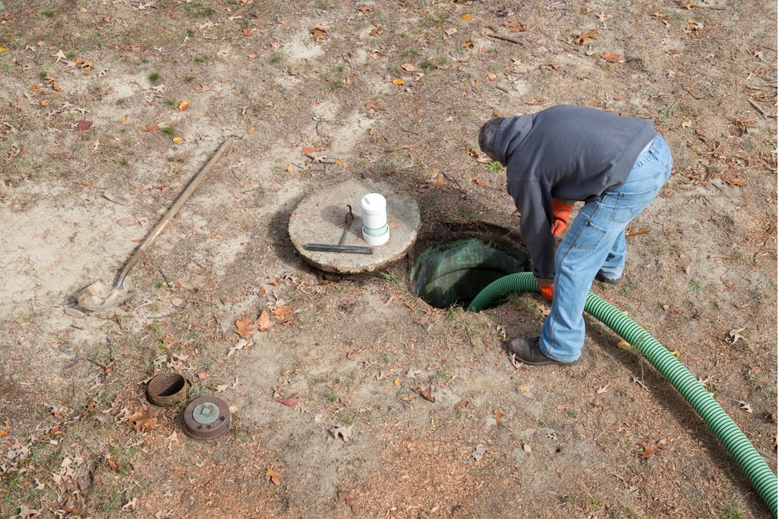 Worker bending over green hose beside manhole cover on dirt ground