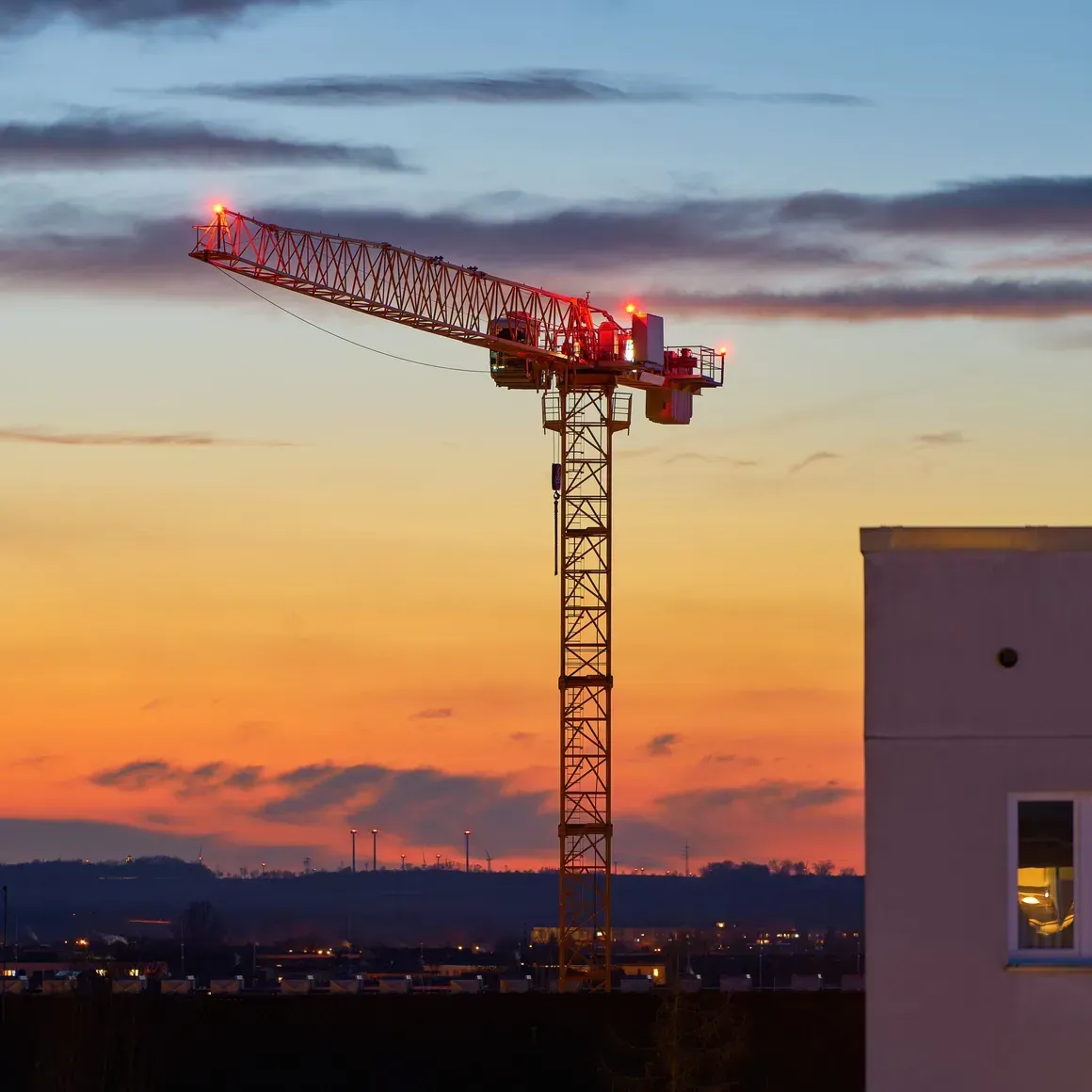 Construction crane lit at dusk against a colorful sunset sky, beside a building silhouette