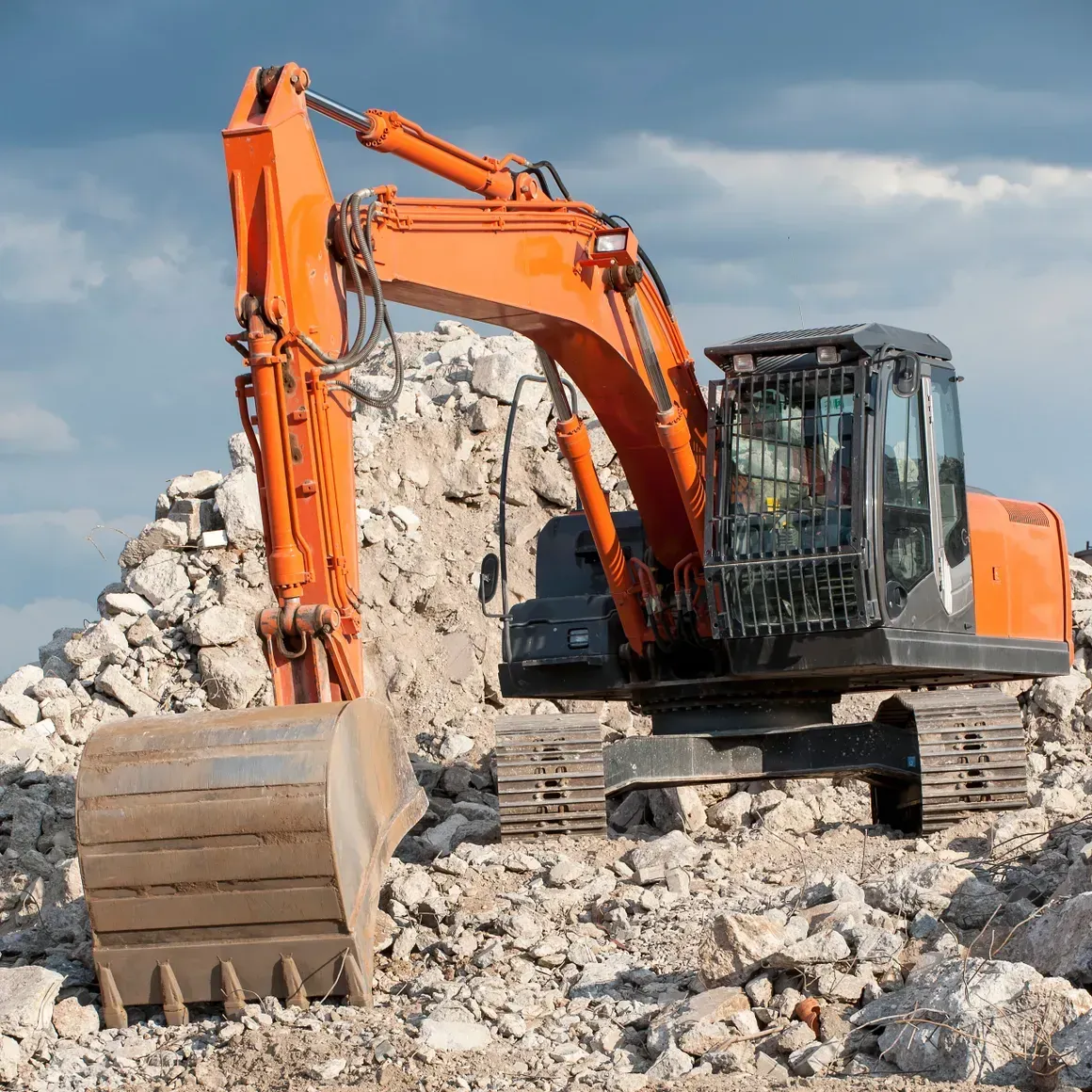 Orange excavator at a rubble site, scooping debris against a cloudy sky.