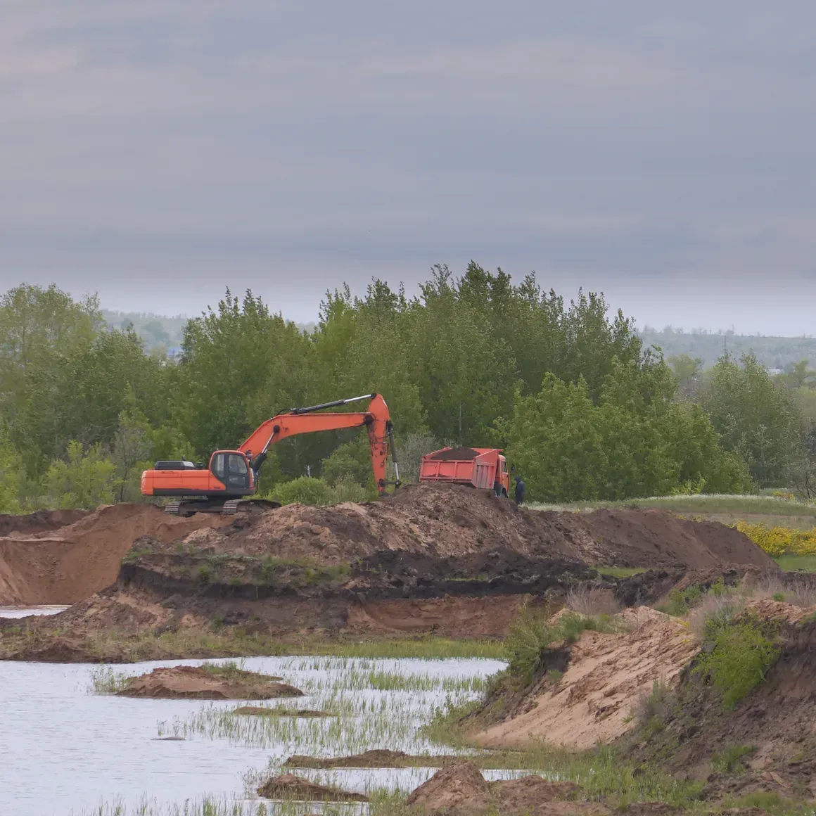 Two orange excavators on a muddy riverbank beside water and trees
