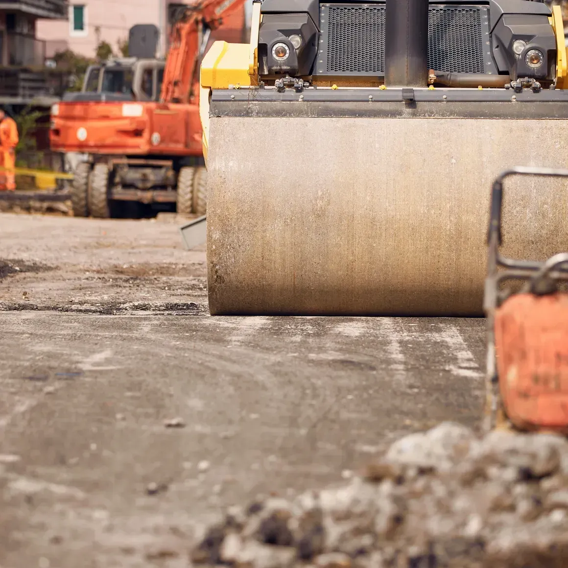 Heavy construction roller compacting a dirt road, with excavators in the background