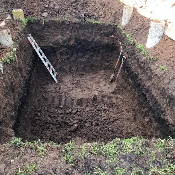 Excavated rectangular pit in dirt, with a ladder leaning inside and stakes marking the edges.