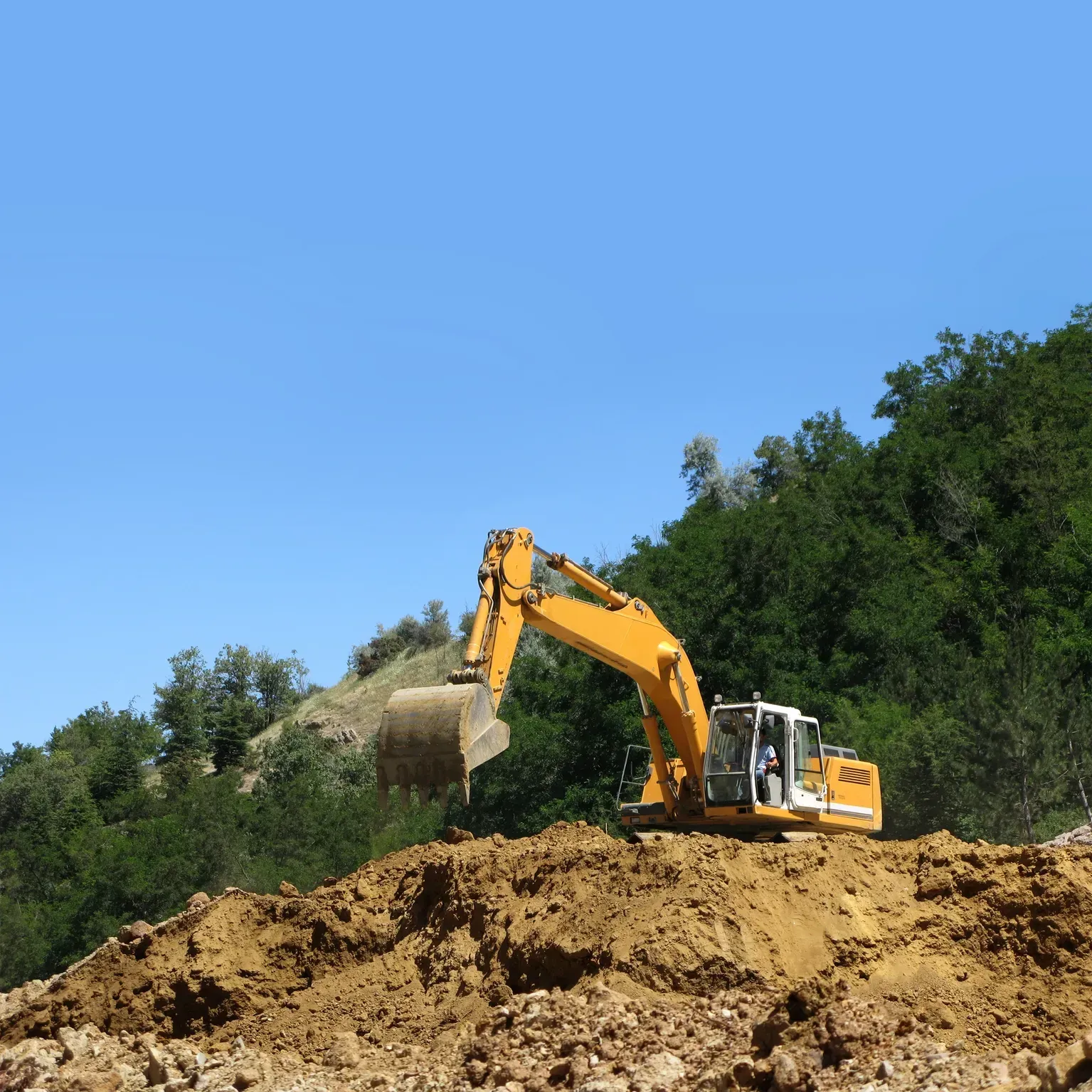 Yellow excavator on a dirt mound under a blue sky, with trees in the background.