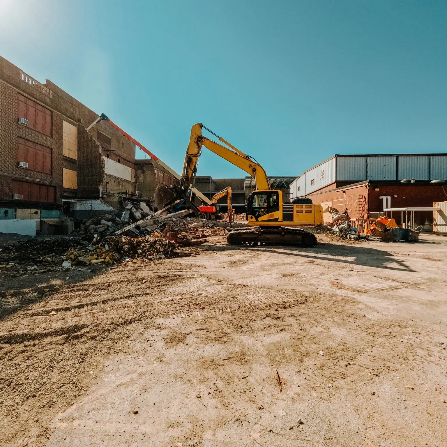 Demolition site with excavator clearing rubble between brick buildings under a blue sky