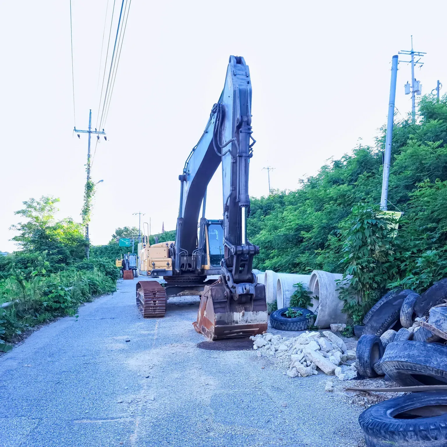 Excavator parked on a gravel roadside beside piles of tires and greenery under utility poles.