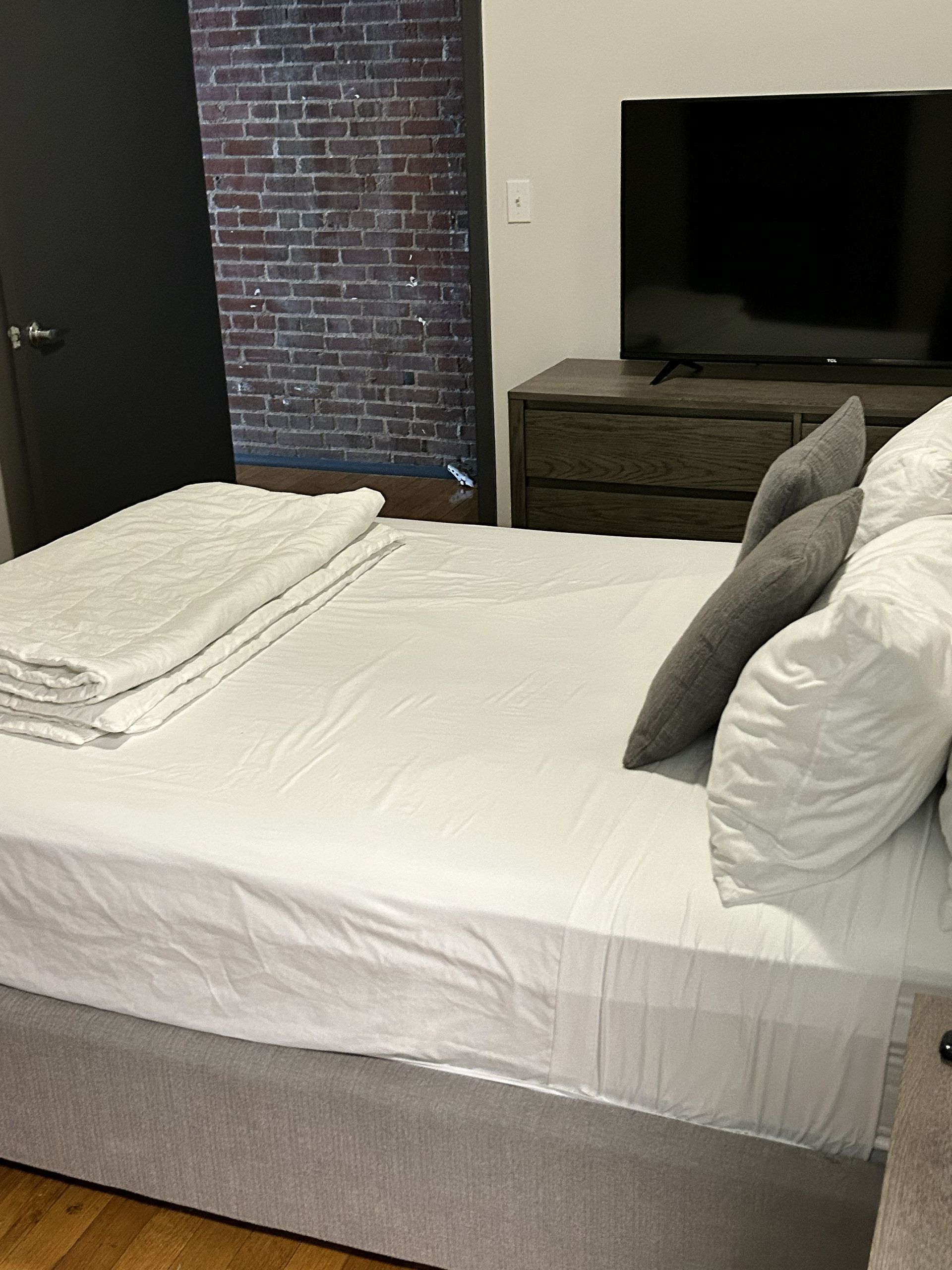 A neatly made bed in a bedroom with white bedding, gray pillows, a brick accent wall, and a TV on a dresser.