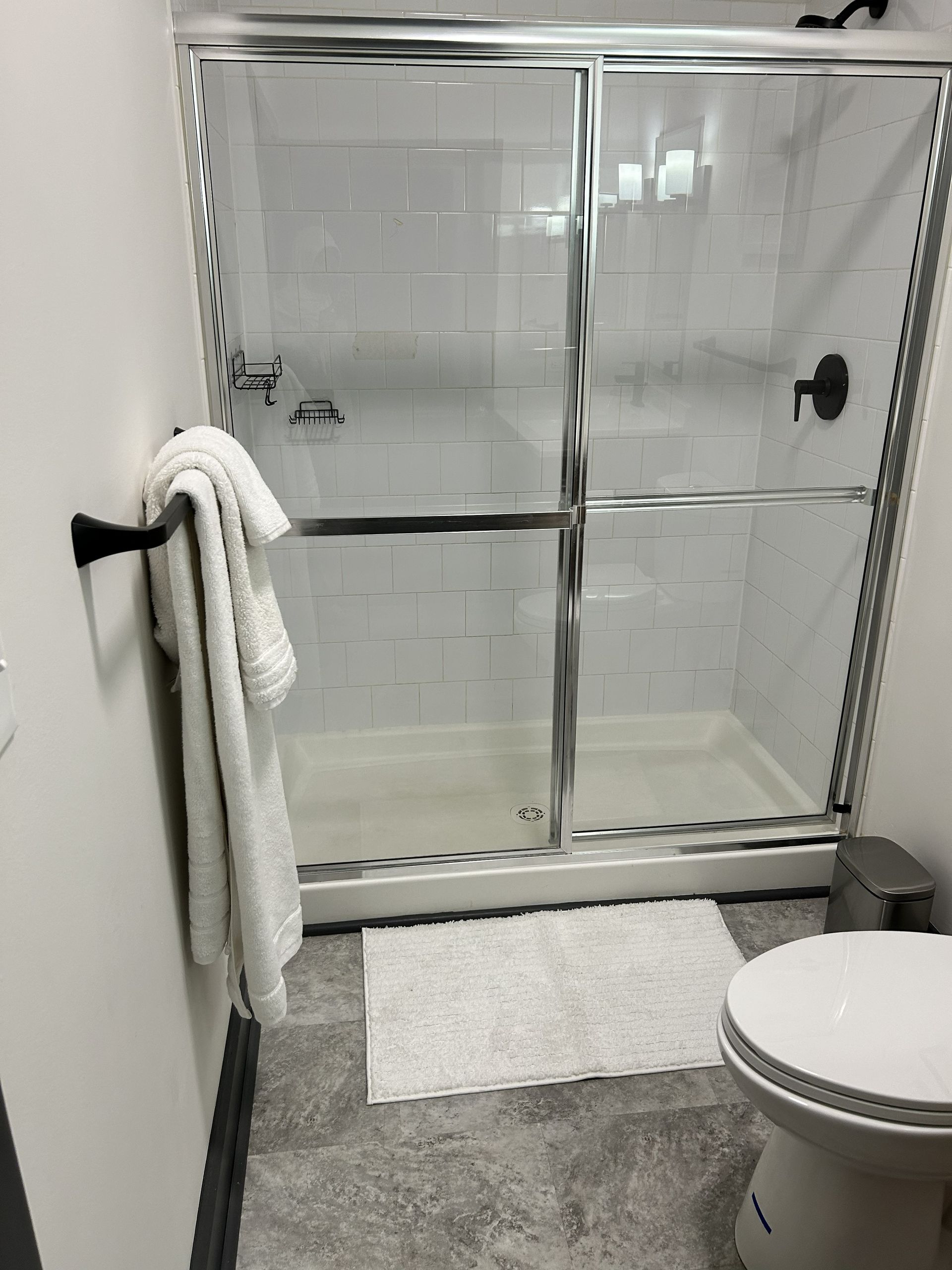 Bathroom with glass shower enclosure, towel on grab bar, bath mat, and partial toilet visible.