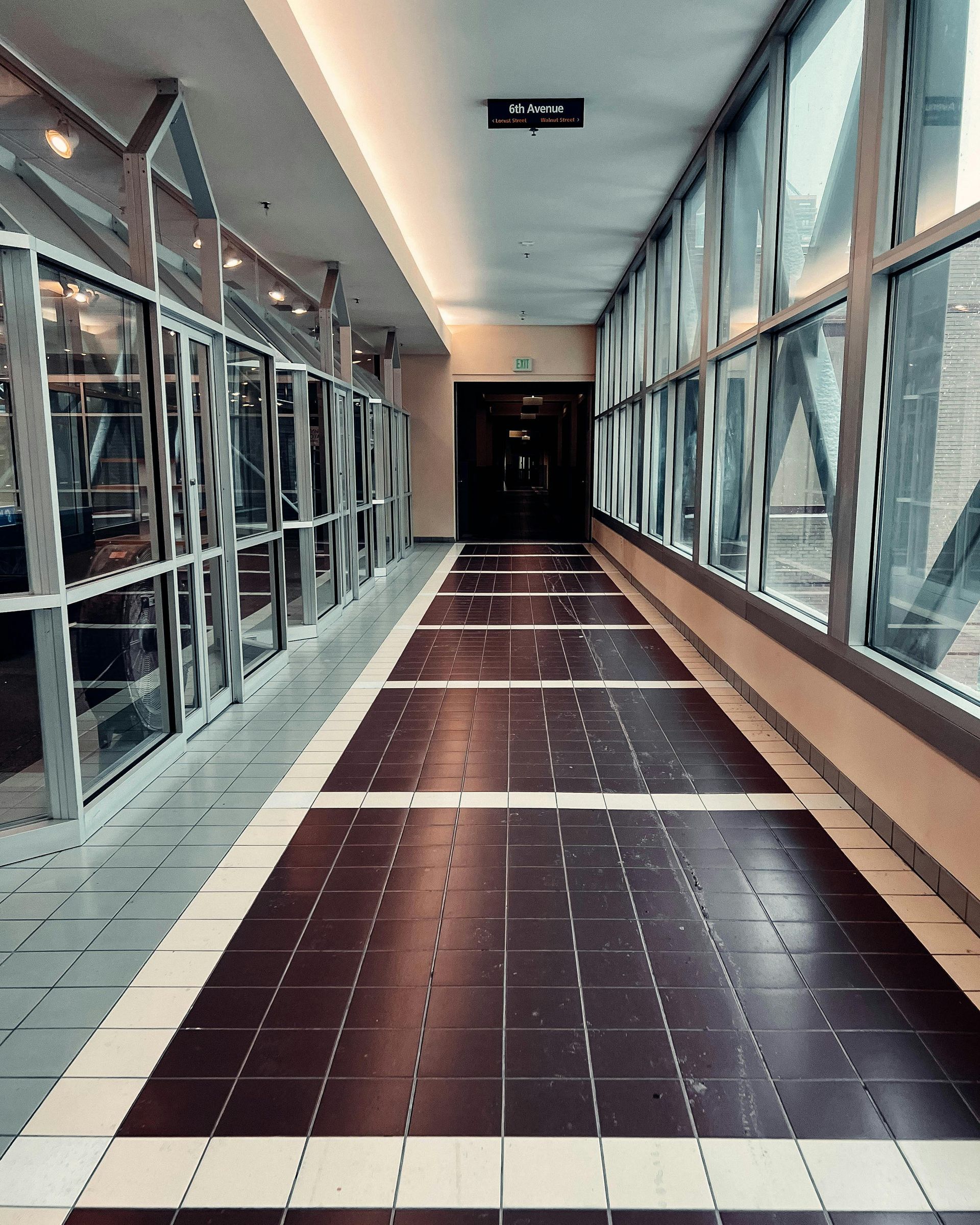 A long, empty indoor corridor with large glass windows on both sides and a dark, tiled walkway leading to a dark doorway.