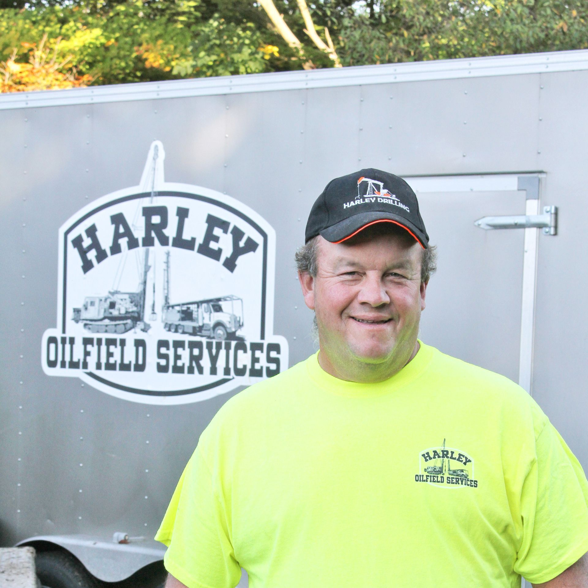 Man in neon shirt and Harley Oilfield Services cap smiles in front of a trailer with the company logo.
