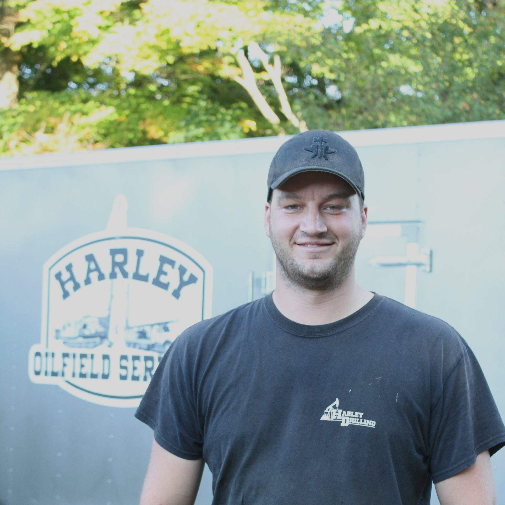 Man in black hat and shirt, smiling in front of a Harley Oilfield Services truck, outdoors.