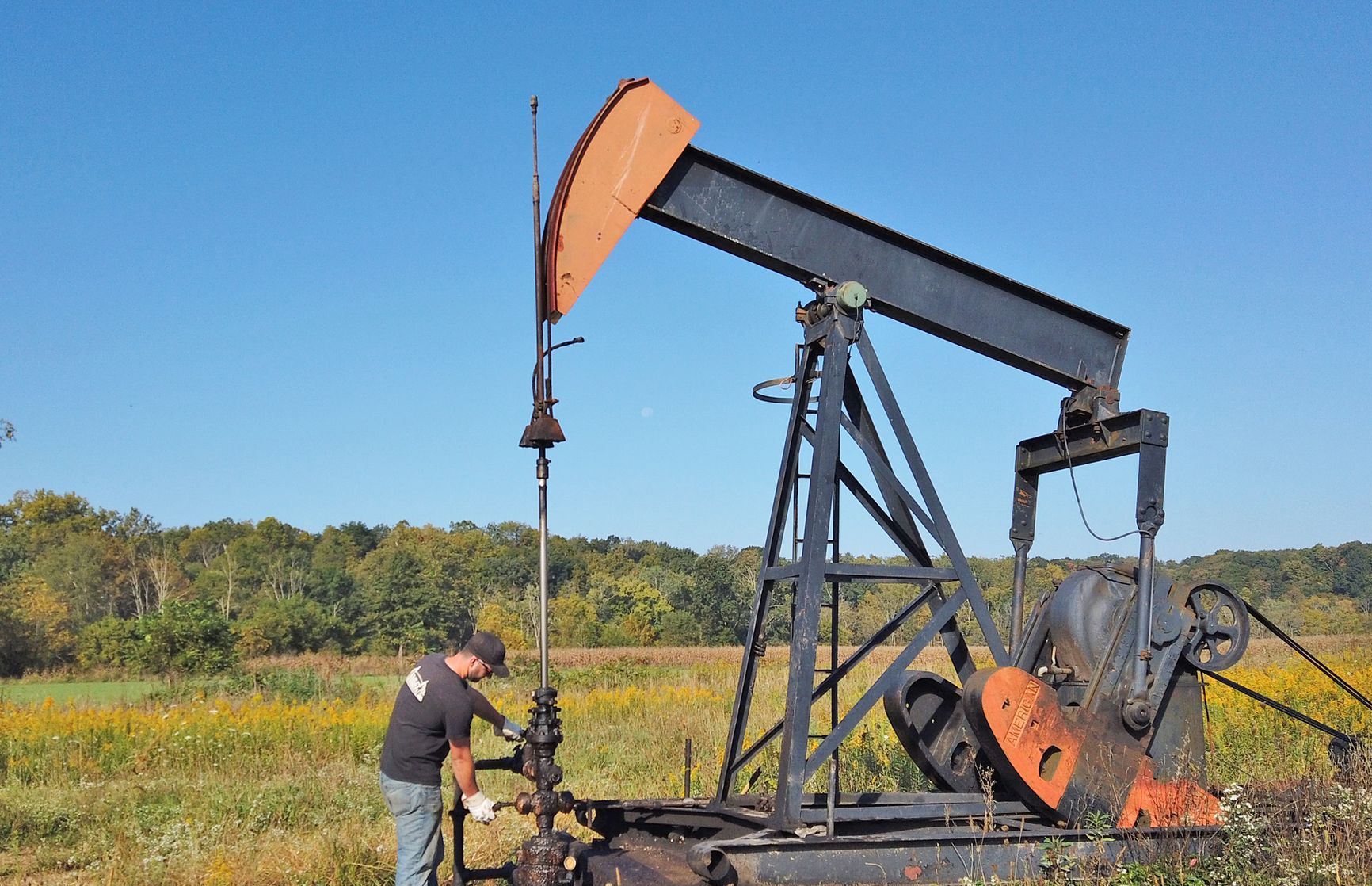 Man working on an oil pump jack in a field under a blue sky.