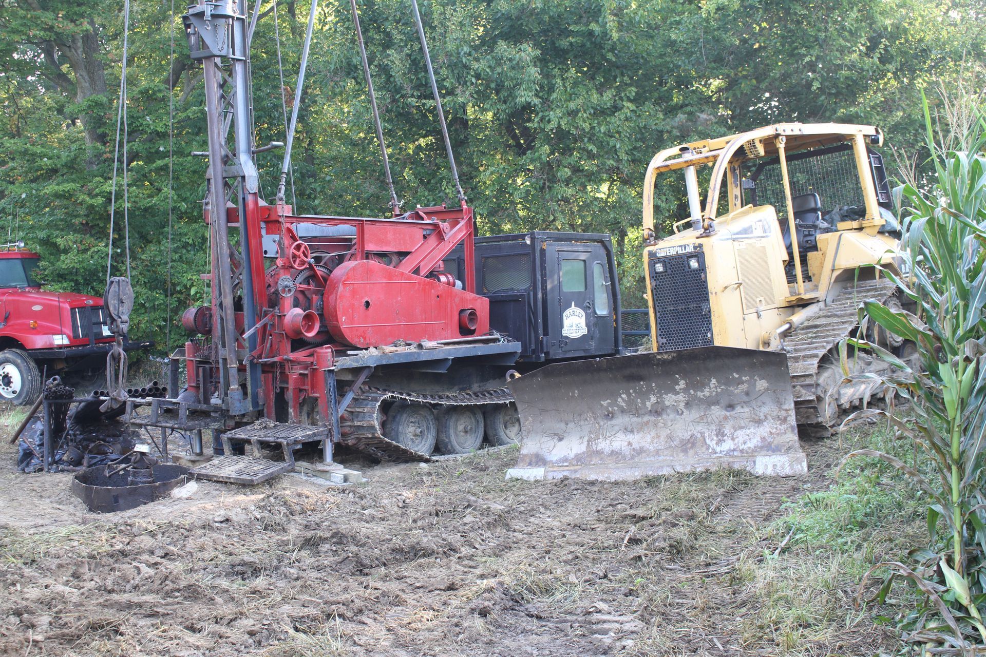 Drilling rig and bulldozer at a work site on muddy ground; red, yellow, green tones.
