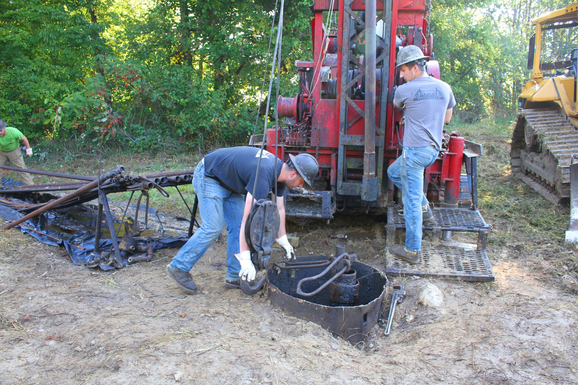 Three workers at a drilling site. One inspects a dark, metal piece while others work near a red machine.