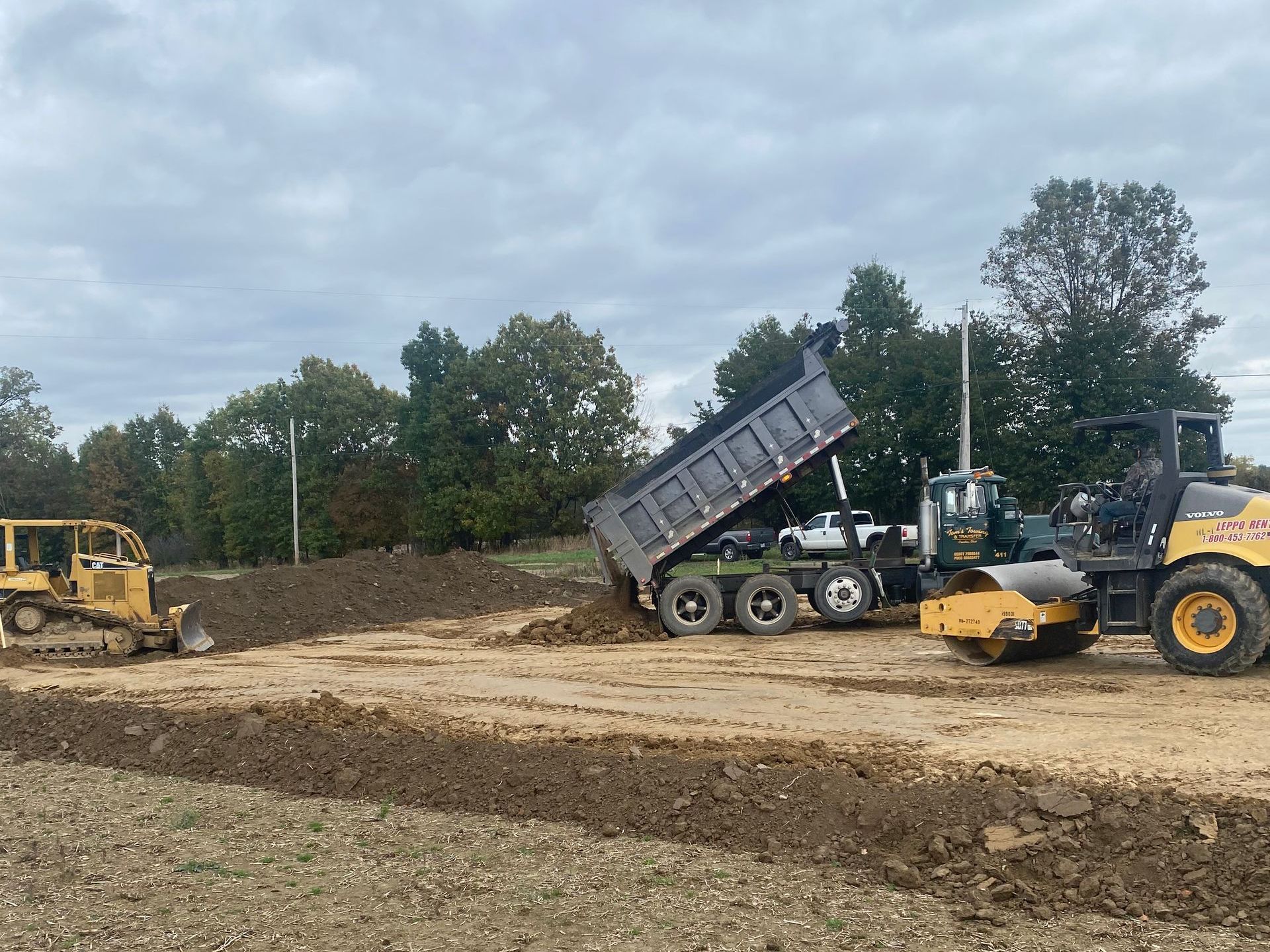Construction site: dump truck unloading soil, bulldozer, and compactor on brown dirt. Trees and cloudy sky.