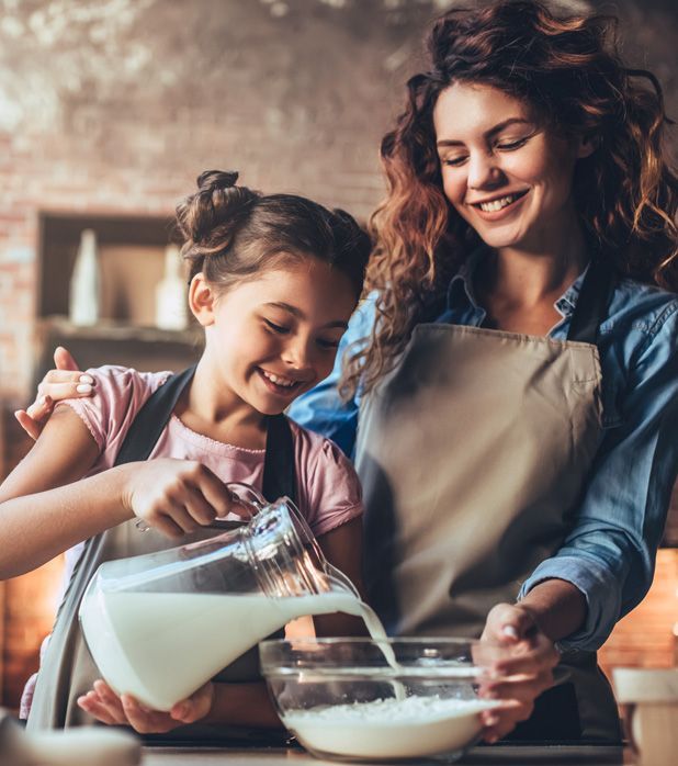 A person and a child wearing aprons smiling while pouring milk from a glass pitcher into a mixing bowl in a kitchen. A person and a child wearing aprons smiling while pouring milk from a glass pitcher into a mixing bowl in a kitchen.