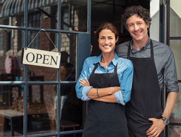 Two people wearing aprons smiling in front of a shop with an Two people wearing aprons smiling in front of a shop with an