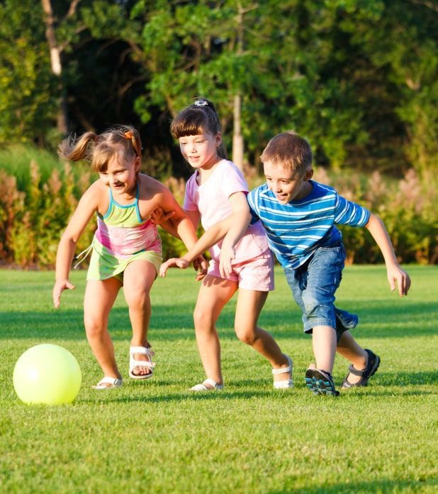 Three children running across a sunlit grass field while chasing a large yellow ball. Three children running across a sunlit grass field while chasing a large yellow ball.
