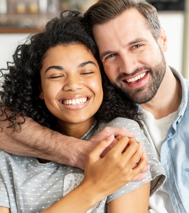 A smiling person with dark curly hair is embraced by another person from behind, both looking happy in a close-up shot. A smiling person with dark curly hair is embraced by another person from behind, both looking happy in a close-up shot.