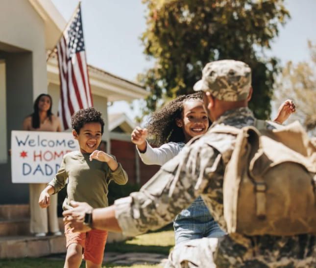 Soldier in uniform hugs two children running towards him, wife holding welcome sign, American flag in background. Soldier in uniform hugs two children running towards him, wife holding welcome sign, American flag in background.