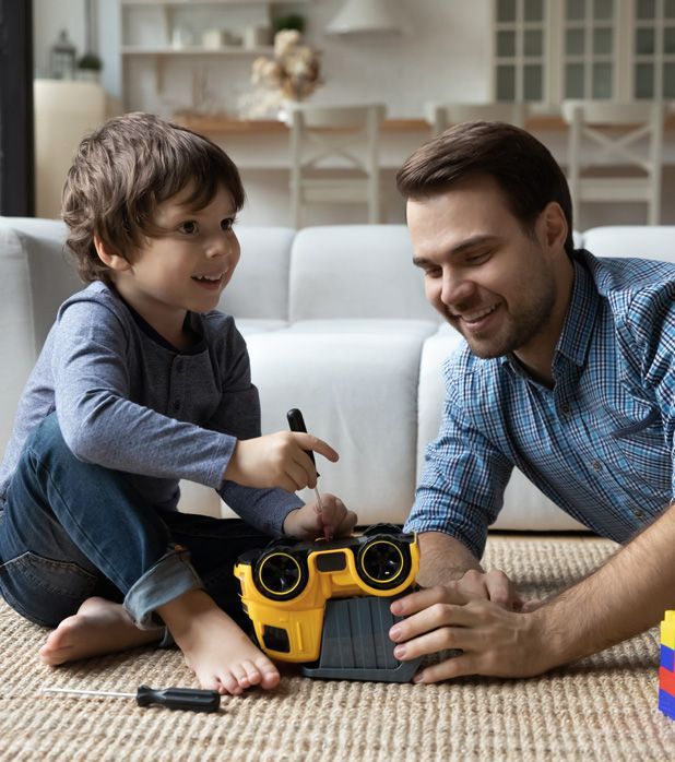 A parent and child sit on a living room rug, smiling as they use a small screwdriver to repair a yellow toy vehicle. A parent and child sit on a living room rug, smiling as they use a small screwdriver to repair a yellow toy vehicle.