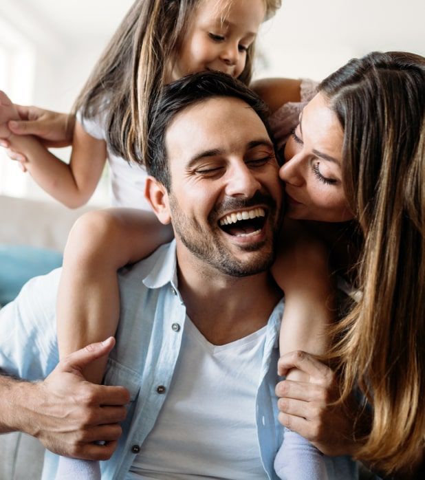 A laughing man gives a child a piggyback ride while a woman kisses his cheek. A laughing man gives a child a piggyback ride while a woman kisses his cheek.