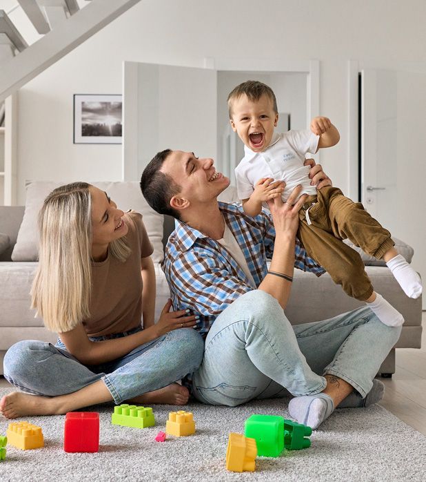A family sits on a living room rug, playing with colorful building blocks as a parent lifts a laughing toddler. A family sits on a living room rug, playing with colorful building blocks as a parent lifts a laughing toddler.