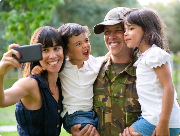 Family taking a selfie; father in camouflage, mother holding phone, children smiling outdoors. Family taking a selfie; father in camouflage, mother holding phone, children smiling outdoors.