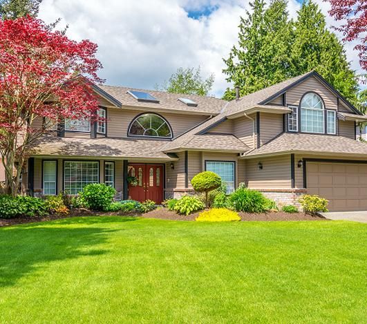 Tan two-story house with red door, large green lawn, and colorful landscaping. Tan two-story house with red door, large green lawn, and colorful landscaping.