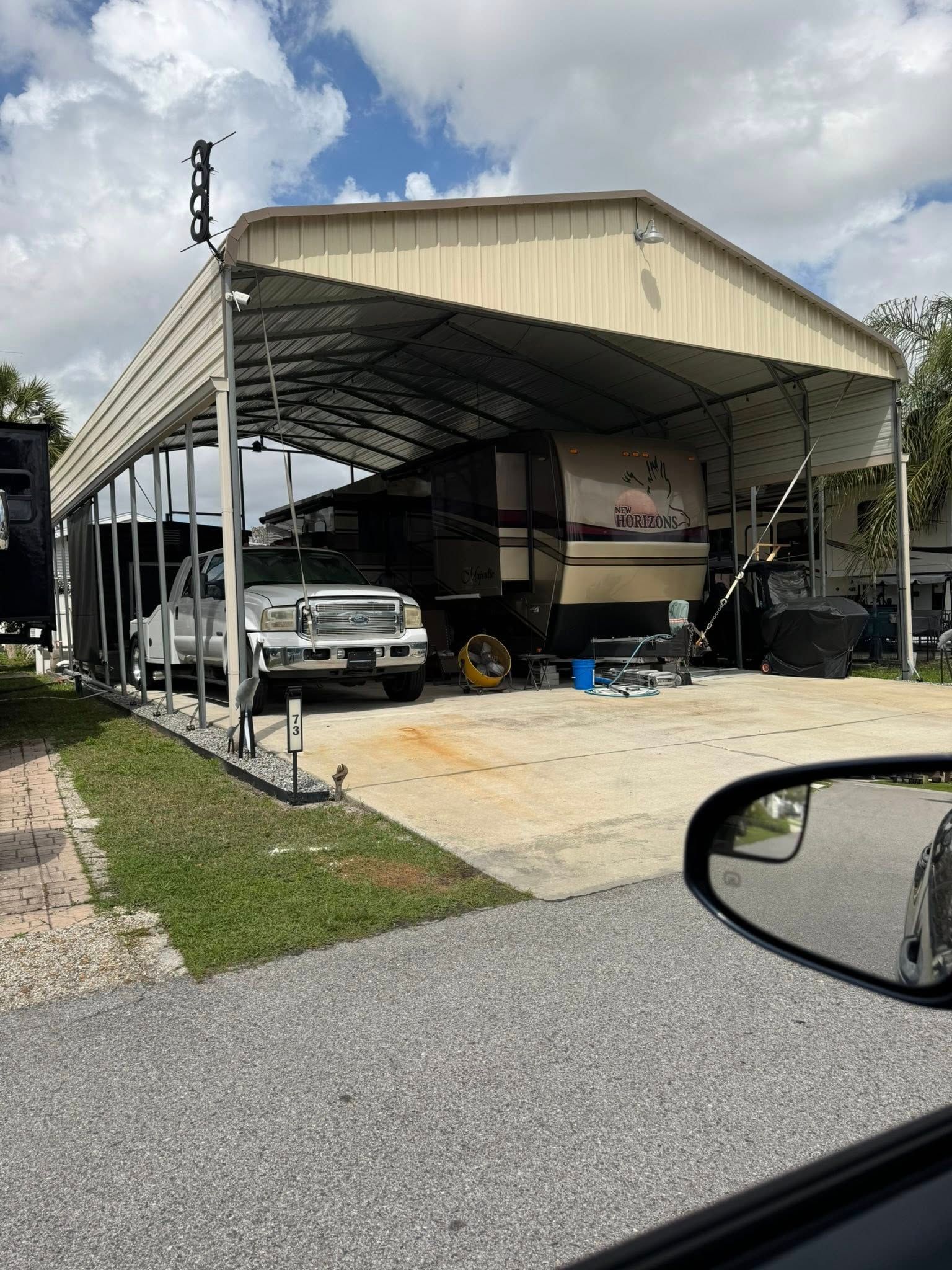 Carport with a white pickup truck and RV parked on a driveway beside a house under a cloudy sky