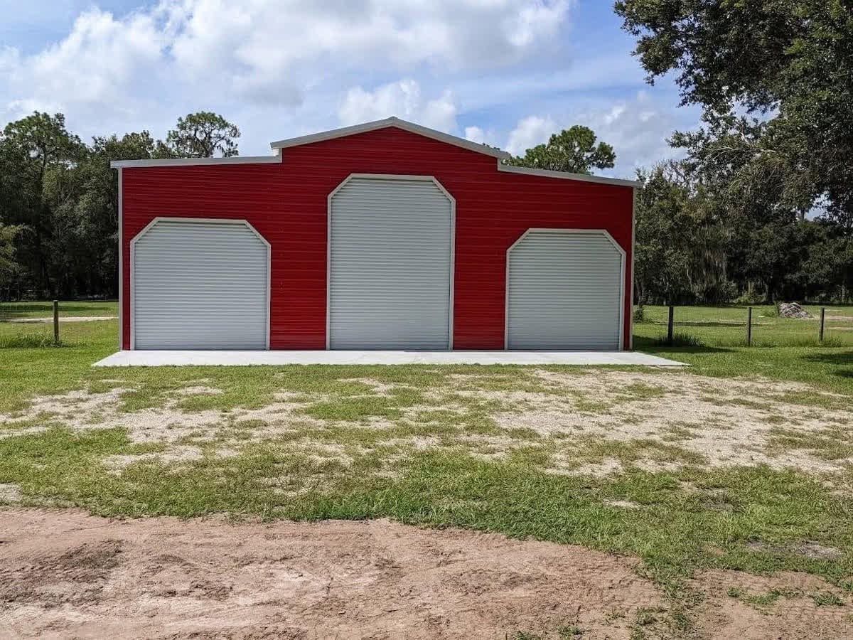 Red barn-style building with three white roll-up doors on a grassy lot under a cloudy sky
