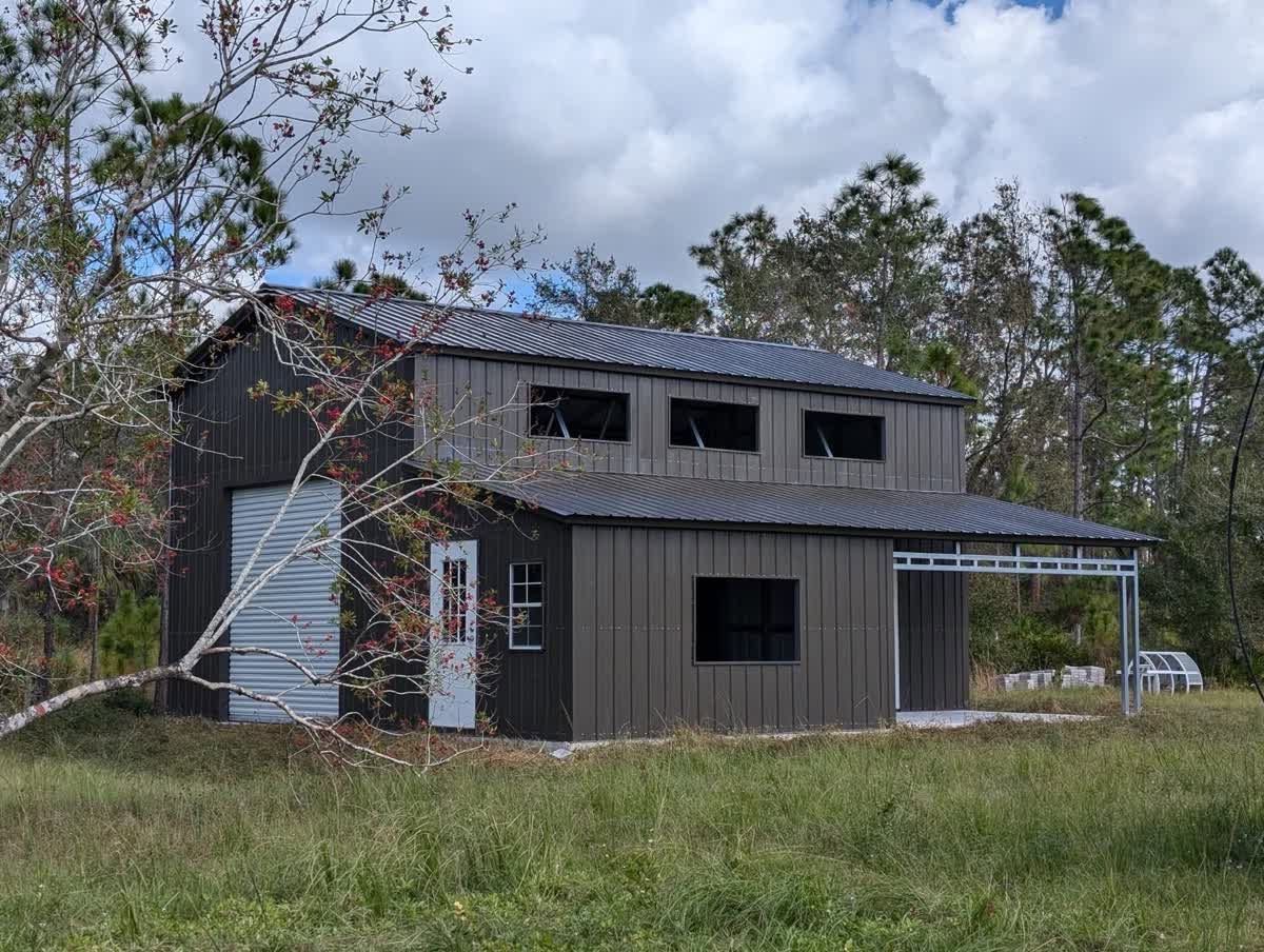 Two-story weathered house with metal roof in a grassy field under cloudy sky