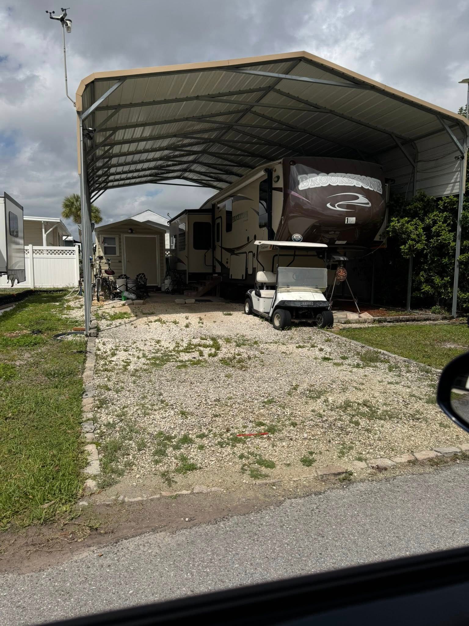 RV parked under a metal carport beside a gravel driveway and road