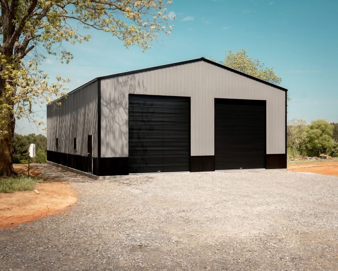 Large white metal barn with black doors on a gravel driveway, surrounded by trees under a clear sky