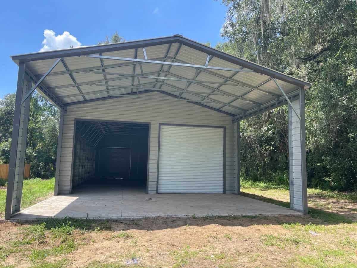 Open-front metal shed with a concrete floor and two roll-up doors, set on a grassy lot under a blue sky