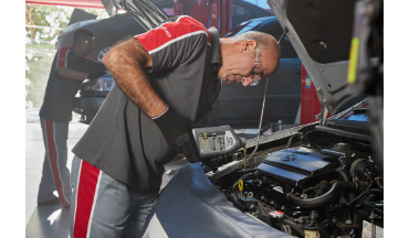 Dois mecânicos trabalhando no motor de um carro em uma oficina. Um deles observa atentamente, usando óculos de segurança.