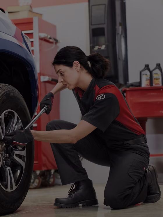 Mecânico ajoelhado, apertando as porcas da roda de um carro. Uniforme vermelho e preto, em uma oficina.