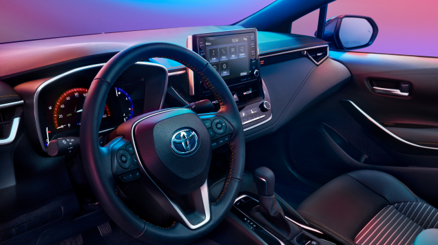 Visão interna de um Toyota Corolla, com foco no volante, painel e tela do sistema de infoentretenimento.