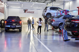 Interior de uma oficina mecânica com carros em elevadores, mecânicos trabalhando e pessoas observando.