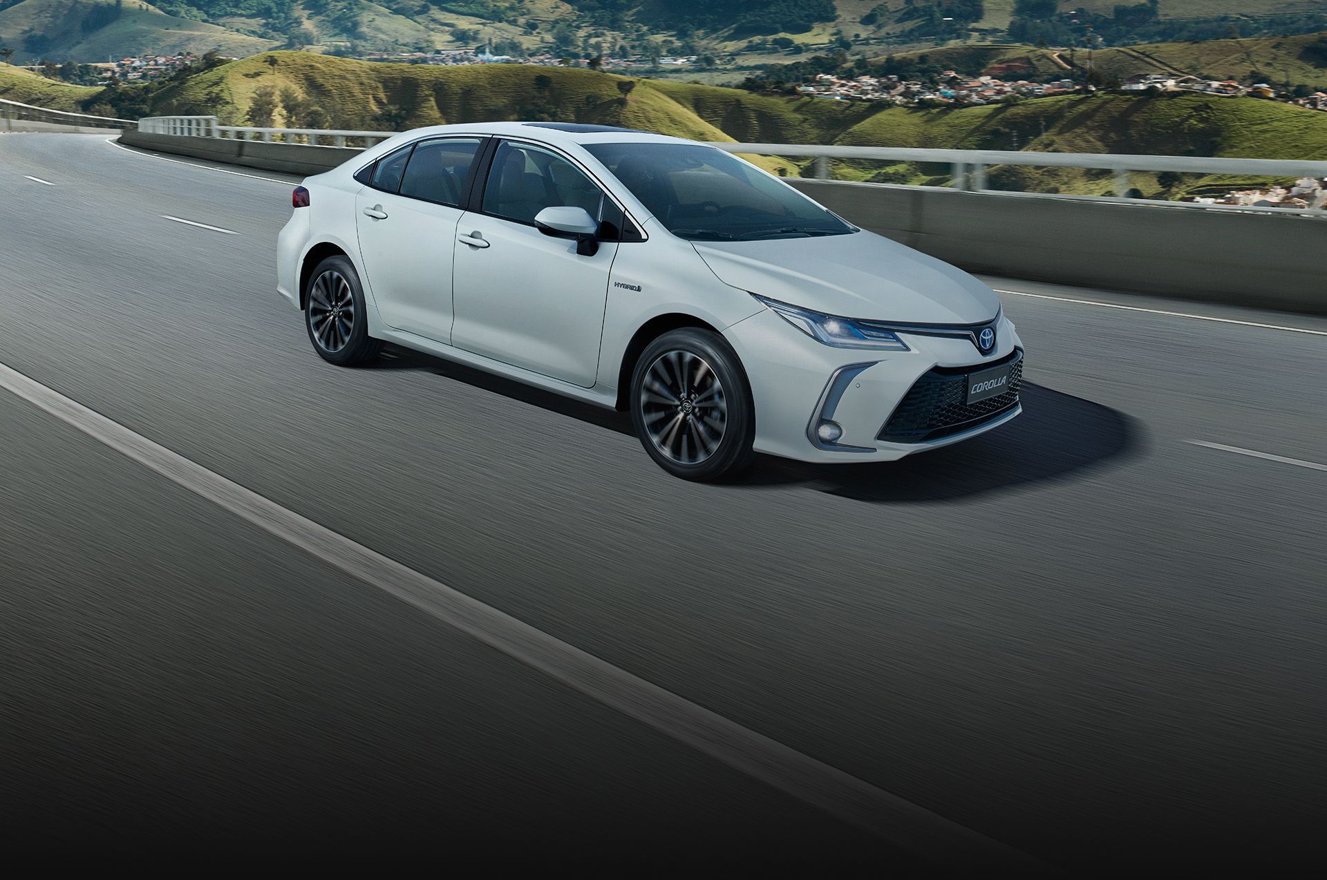 Um Toyota Corolla branco trafegando em uma rodovia, com montanhas ao fundo.
