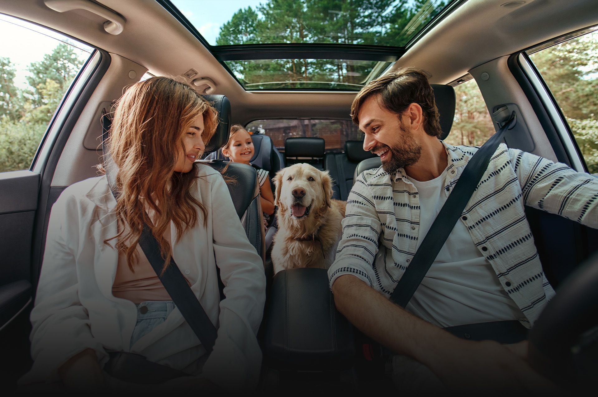 Família e cachorro sorrindo dentro de um carro. A luz do sol entra pelo teto solar; árvores ao fundo.