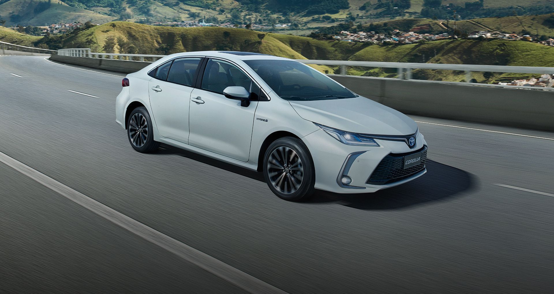 Um Toyota Corolla branco trafegando em uma rodovia, com montanhas ao fundo.