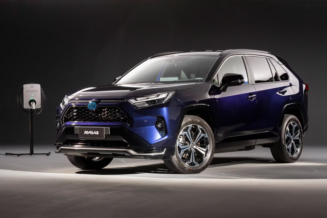SUV Toyota RAV4 Prime azul estacionado ao lado de uma estação de carregamento contra um fundo escuro.