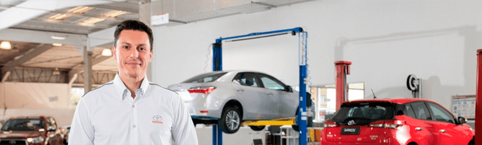 Um mecânico de camisa branca sorri em uma oficina mecânica com dois carros em elevadores.