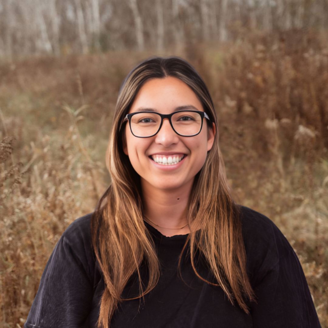 Smiling person with glasses in a black sweater, standing outdoors in a grassy field.