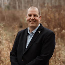Smiling man in a dark blazer standing outdoors against a blurred brown field