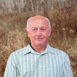 View Bio Portrait of a smiling man in a light blue striped shirt outdoors, with tall grasses behind him