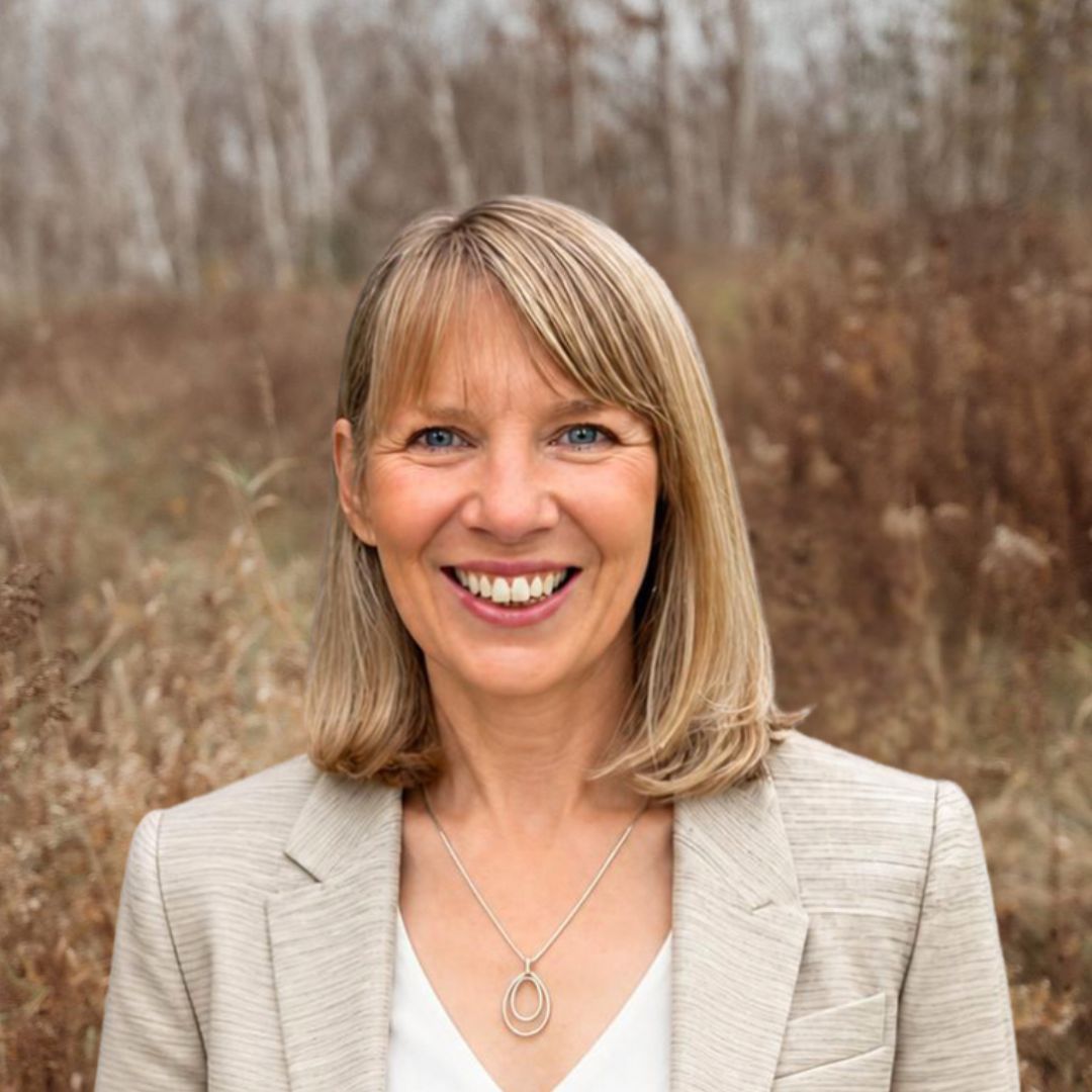 View Bio Smiling woman in a beige blazer outdoors with a blurred brown field and trees behind her